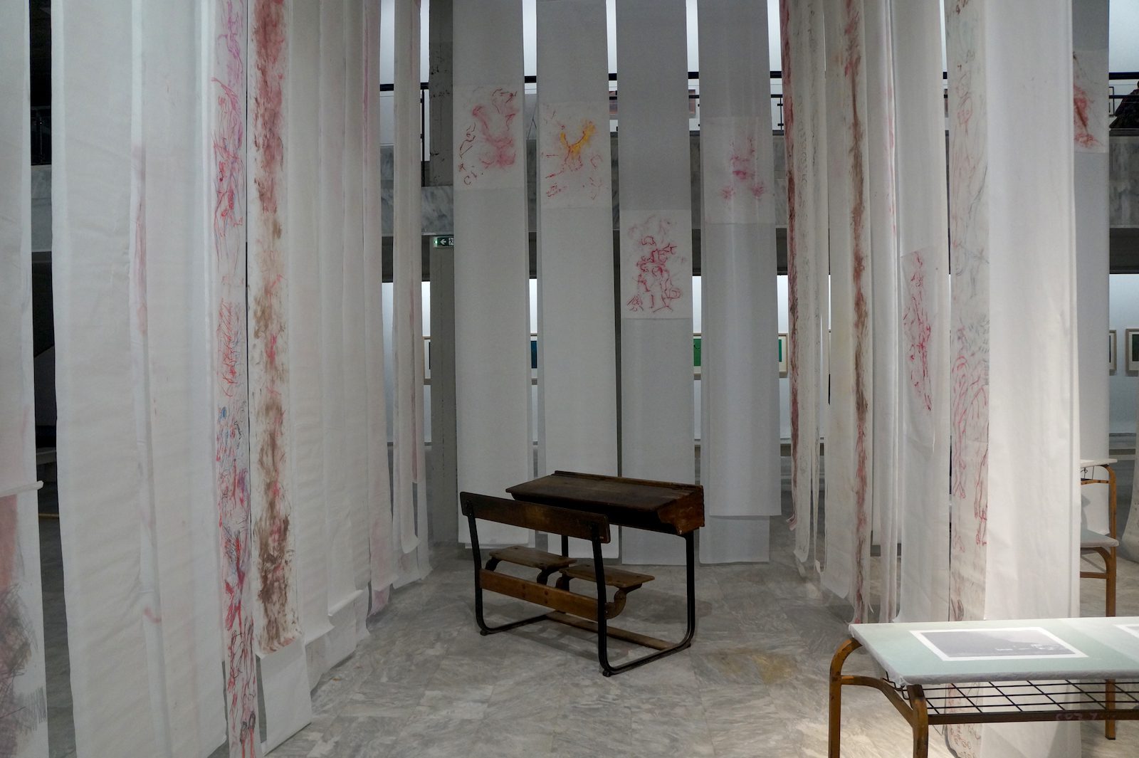 Pélagie Gbaguidi, The Missing link Dicolonisation Education by Mrs Smiling stone, Documenta 14, Athens 2017. Installation, school desks, photographs of Dr. Peter Magubane, glassine paper sheets, drawings with color pencil, earth and lipstick on transparent paper, video, dimensions variable. Courtesy of the artist.